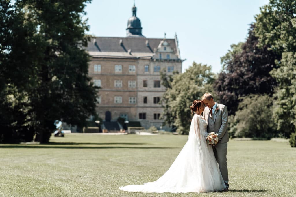 Brautpaarshooting vor dem Schloss Wolfsburg auf der Wiese: Braut und Br&auml;utigam stehen Stirn an Stirn und halten gemeinsam den Blumenstrau&szlig;