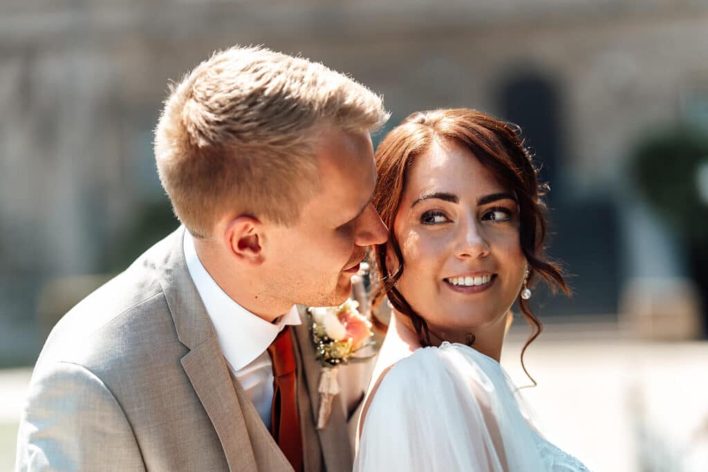 Brautpaarshooting am Schloss Wolfsburg: Br&auml;utigam fl&uuml;stert der Braut etwas ins Ohr, Braut reagiert l&auml;chelnd, intimer Moment