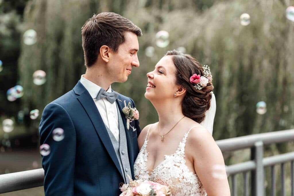 Brautpaarshooting im Schlosspark Braunschweig: Brautpaar steht auf einer Br&uuml;cke, schaut sich l&auml;chelnd an, Seifenblasen im Hintergrund