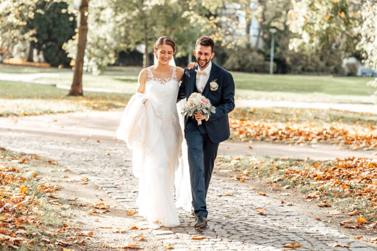Brautpaarshooting in Fallersleben: Brautpaar l&auml;uft gemeinsam durch Herbstlaub, Br&auml;utigam h&auml;lt den Blumenstrau&szlig;, beide lachen herzlich