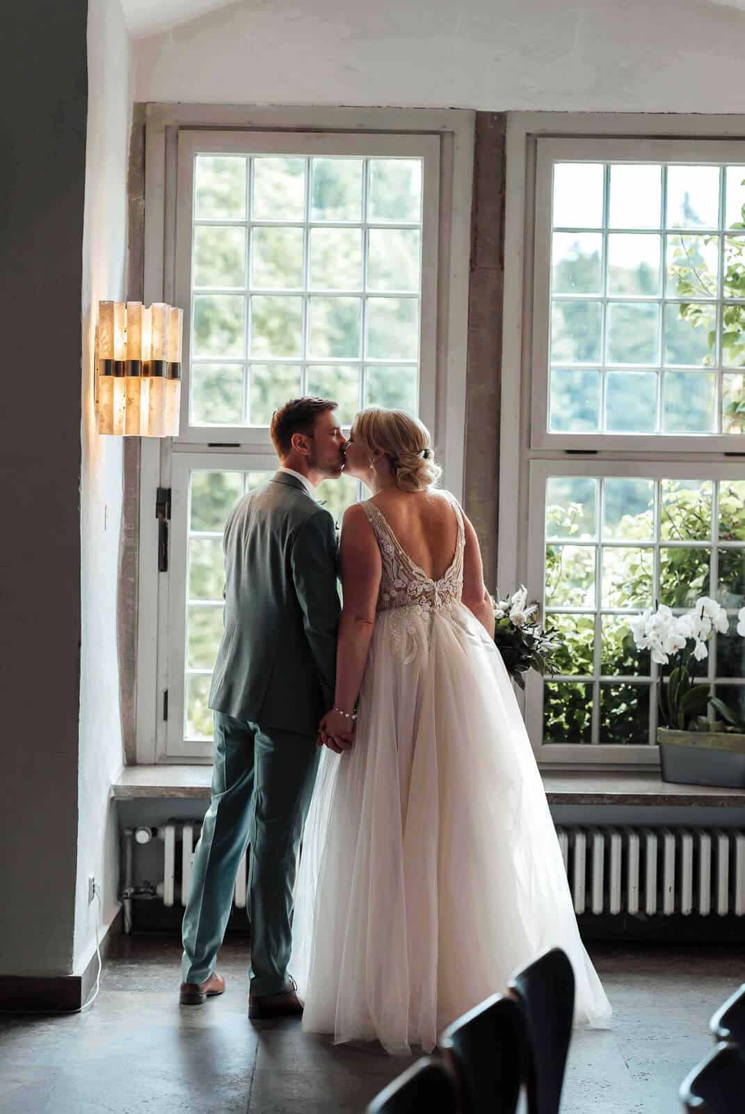 Brautpaar küsst sich im Schloss Wolfsburg vor einem großen Fenster, romantischer Moment in eleganter Kulisse
