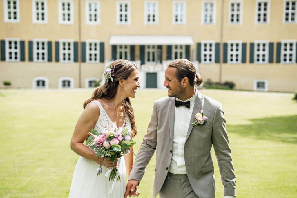 Brautpaarshooting am Schloss Destedt auf der Wiese: Brautpaar hält sich die Hände und lächelt sich an, natürlicher Moment