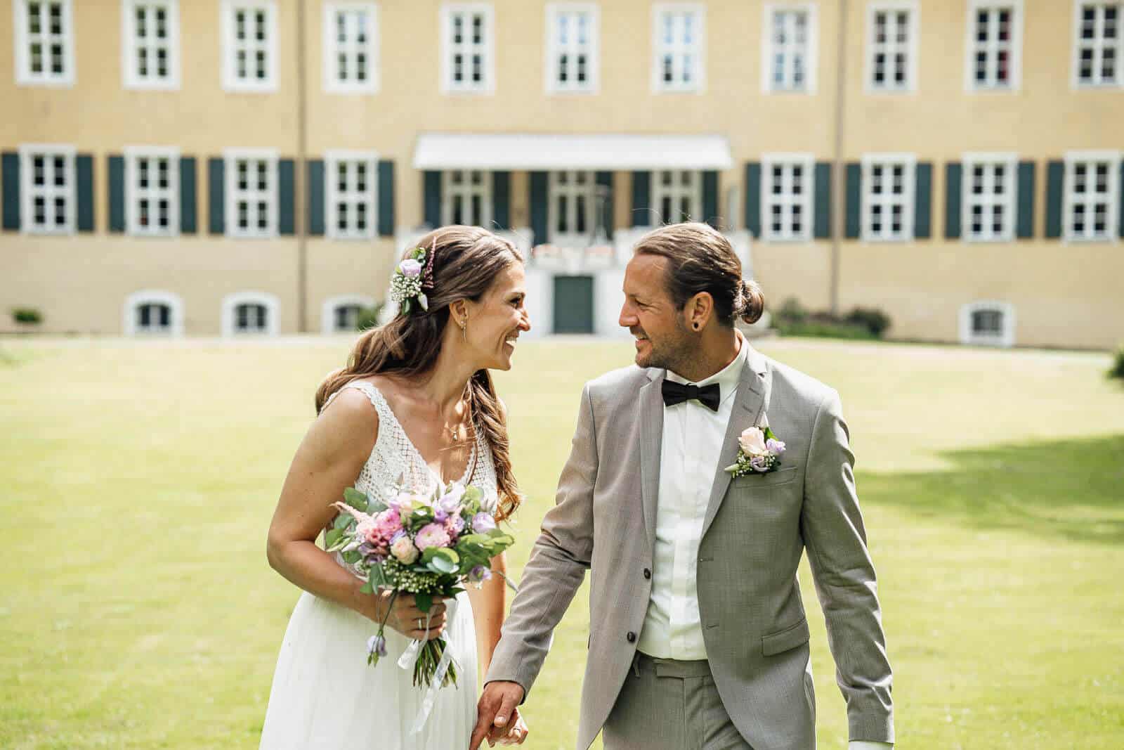 Brautpaarshooting am Schloss Destedt auf der Wiese: Brautpaar hält sich die Hände und lächelt sich an, natürlicher Moment