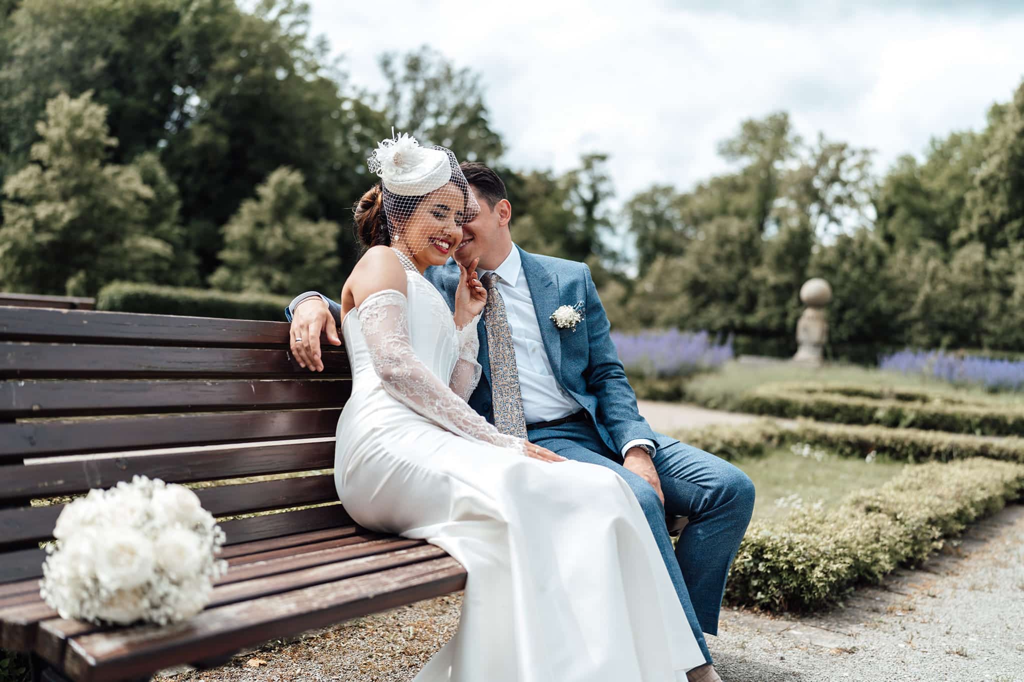 Brautpaarshooting im Park: Braut trägt einen eleganten Hut, Brautpaar sitzt gemeinsam auf einer Bank, stilvoller Moment