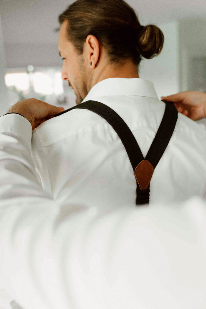 Bräutigam macht sich zuhause fertig und trägt braune Hosenträger beim Getting Ready, natürlicher Vorbereitungsmoment vor der Hochzeit