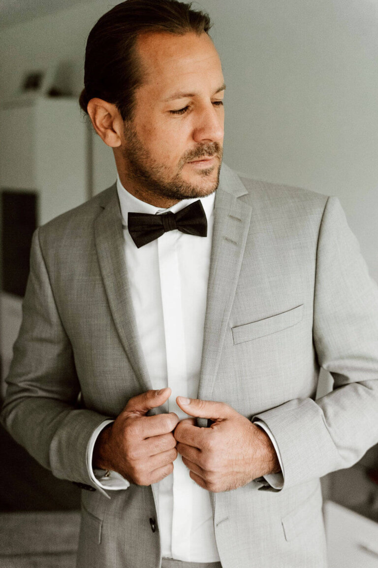 Bräutigam beim Getting Ready zuhause am Fenster trägt Krawatte und grauen Anzug, ruhiger Vorbereitungsmoment vor der Hochzeit