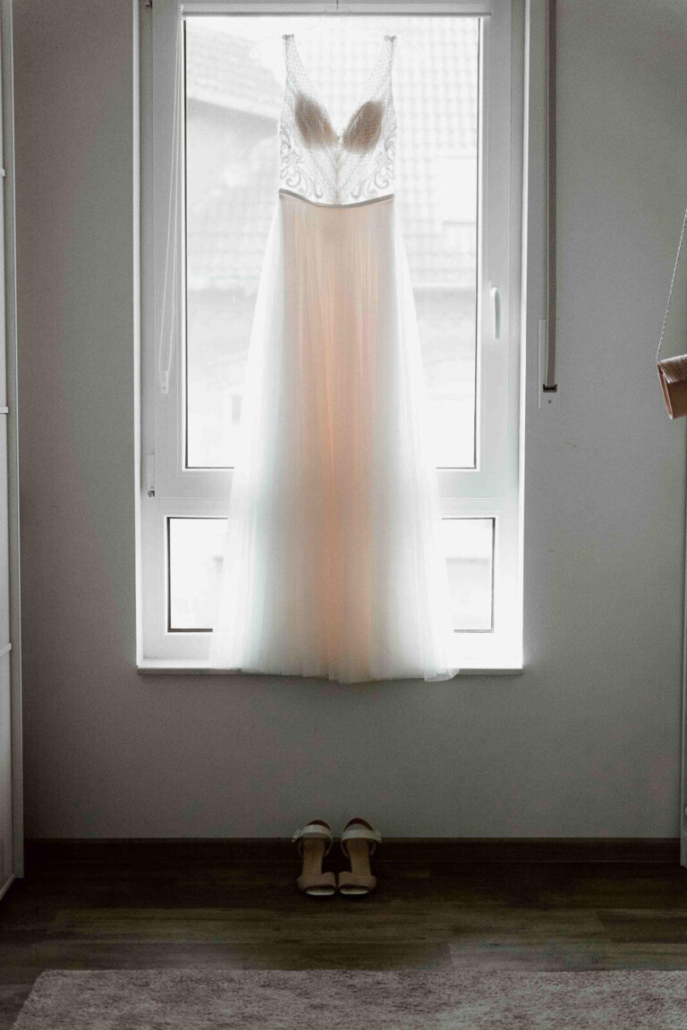 Brautkleid hängt beim Getting Ready zuhause am Fenster, Detailaufnahme im natürlichen Fensterlicht
