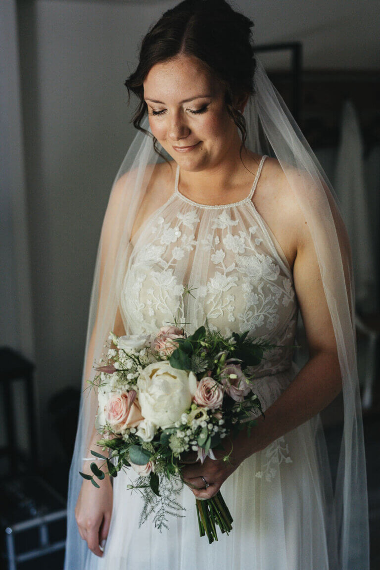 Getting Ready der Braut in Heese 5: Braut steht am Fenster und schaut ihren Brautstrauß an, ruhiger Moment im natürlichen Licht