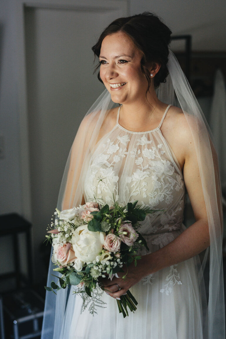 Braut beim Getting Ready in Heese 5 steht am Fenster mit Blumenstrauß in der Hand und trägt ein Brautkleid mit Schleppe