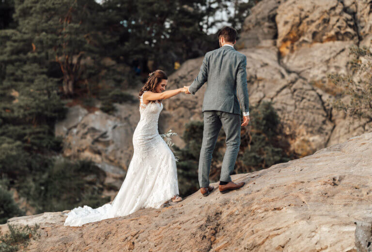 Braut und Bräutigam stehen auf einem Felsen, Bräutigam hilft der Braut nachzukommen, romantischer Moment in der Natur