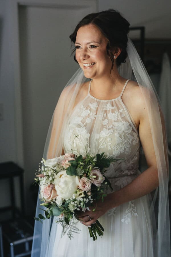 Hochzeitsfotograf Wolfsburg – Braut hält Brautstrauß und lächelt beim Getting Ready am Fenster